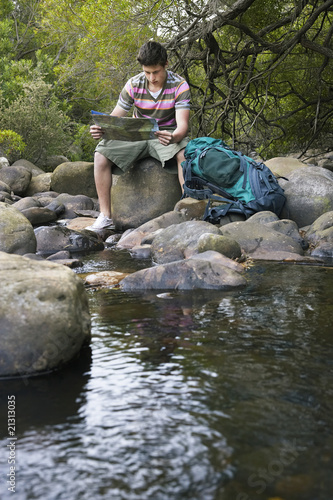 teenage boy (16-17 years) sitting on stone by river reading map