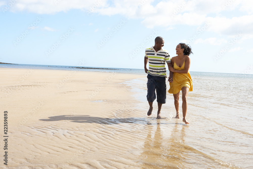 Romantic Young Couple Walking Along Shoreline Of Beach Holding H