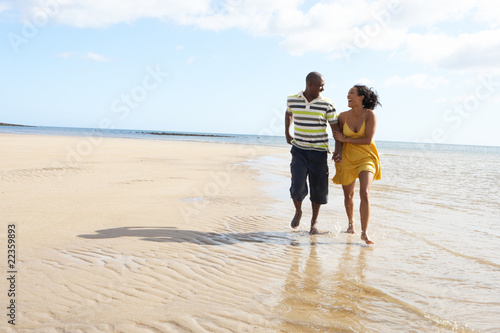Romantic Young Couple Walking Along Shoreline Of Beach Holding H