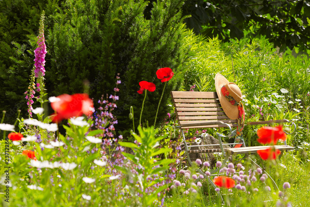 garden bench with straw hat within summer flowers 01 - obrazy, fototapety, plakaty 