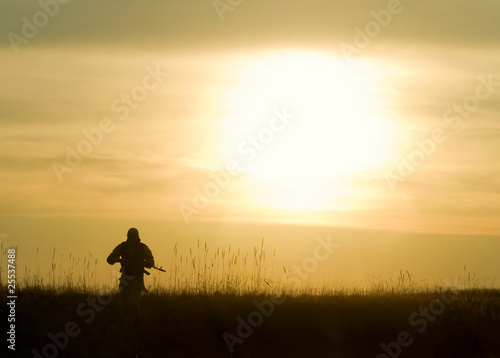 Man with rifle walking on grassy landscape during sunset