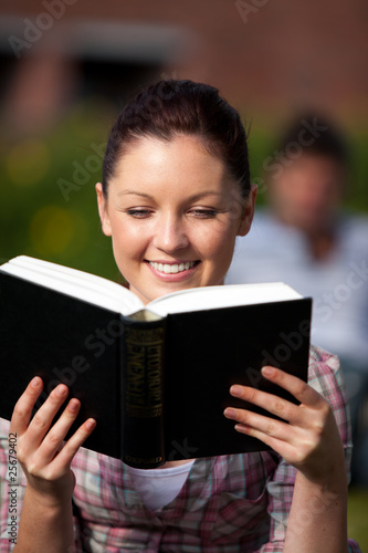 Smiling female student reading a book sitting on grass at