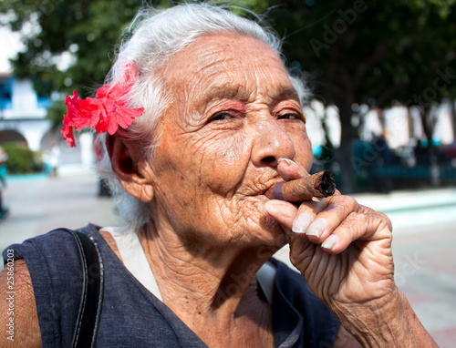 Photography Old wrinkled woman with red flower smoking cigar