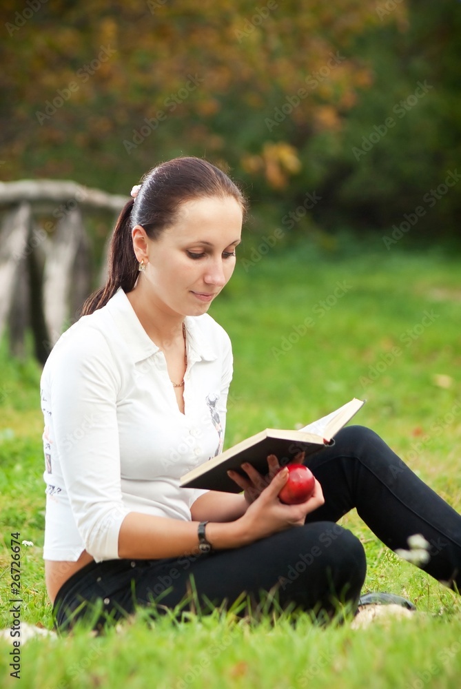 young woman sitting on grass with apple and book