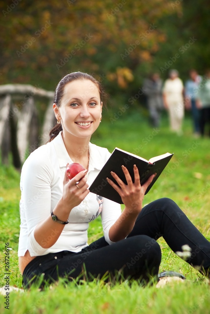 youn smiling woman sitting on green grass with book and apple