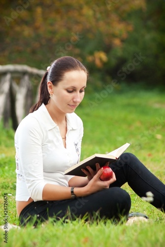 young woman sitting on grass with apple and book