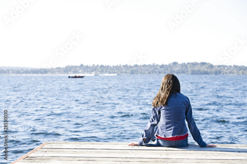 Girl sitting alone on dock by lake, looking out over water.