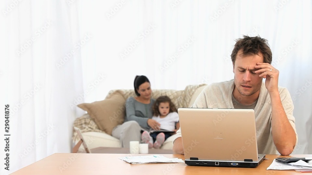 Angry man on his laptop while his family is on a sofa Angry man on his laptop while his family is on a sofa