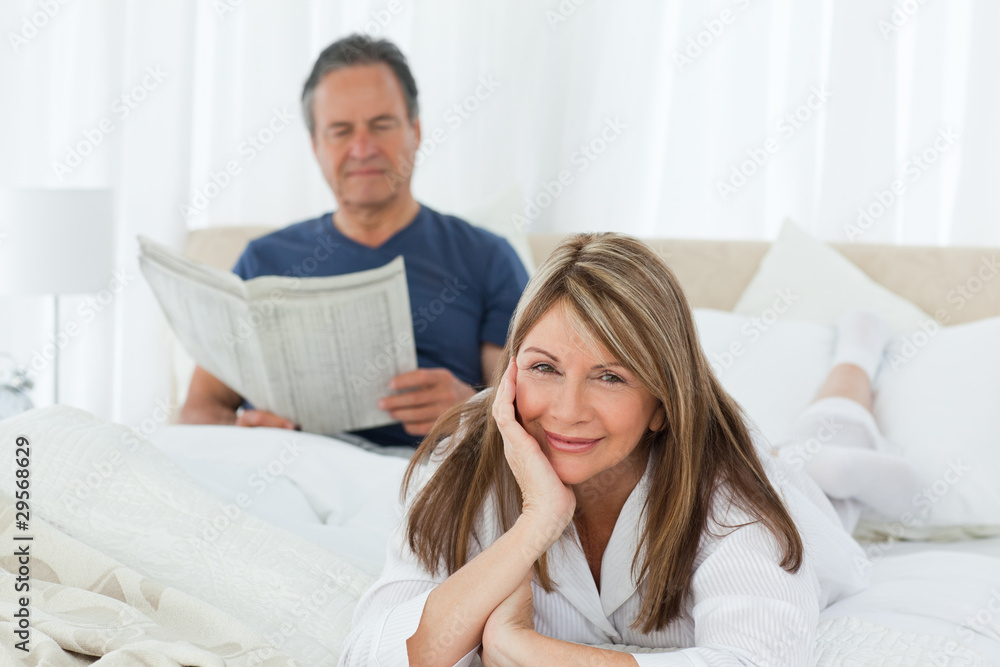 Smiling woman looking at the camera while her husband is readin Smiling woman looking at the camera while her husband is readin