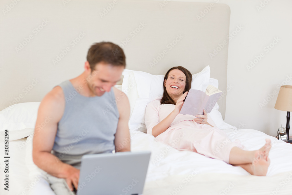 Manworking on his laptop while his wife is reading a book on the Manworking on his laptop while his wife is reading a book on the