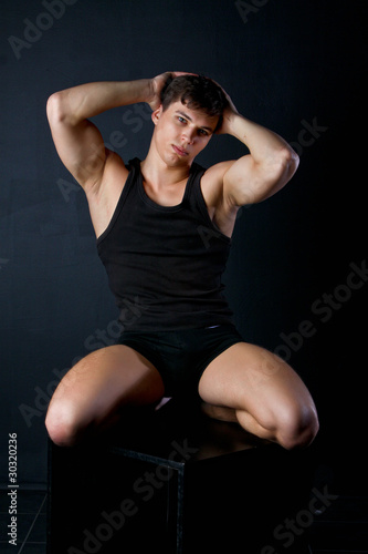 Muscle young man sits in black clothes on a black cube in dark