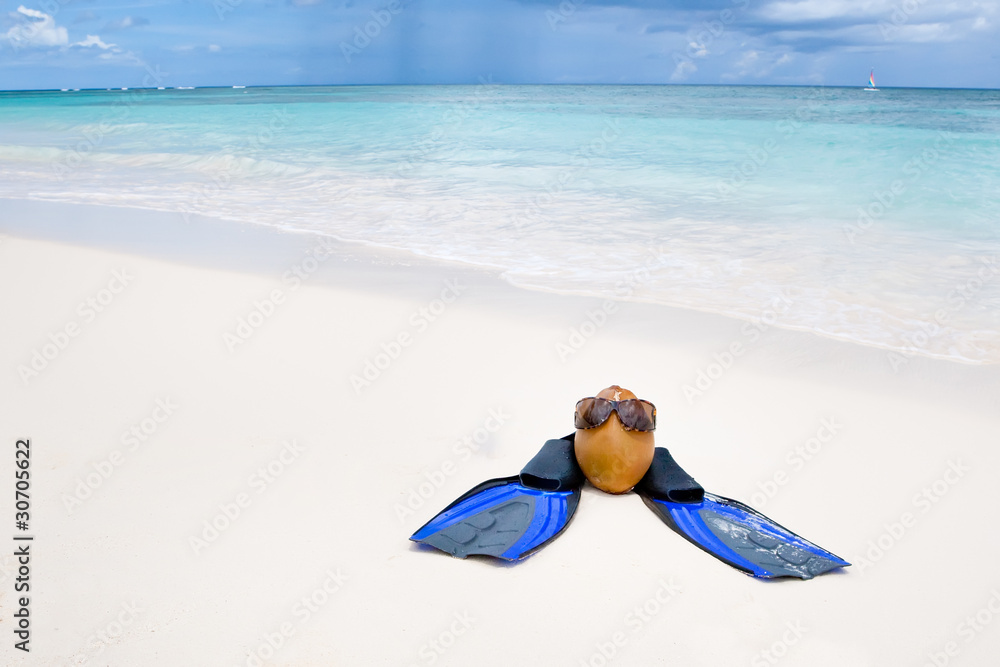 Coconut with black sunglasses and blue flippers on white sand be