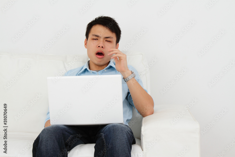 A man yawning while using a laptop sitting on a sofa
