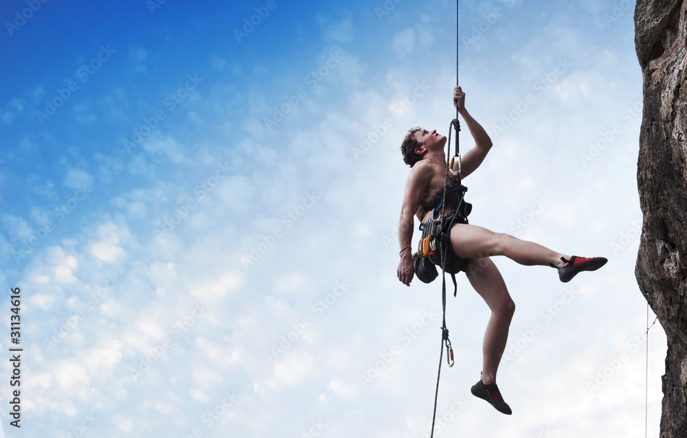 Young male climber hanging on a rope and looking to somewhere on blue cloudy sky background Young male climber hanging on a rope and looking to somewhere on blue cloudy sky background