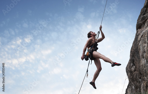 Young male climber hanging on a rope and looking to somewhere on blue cloudy sky background