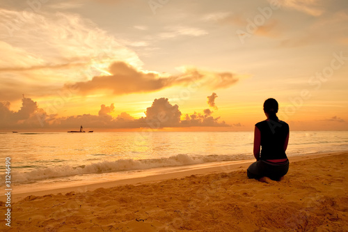 Woman sitting on a sand beach in front of sunset and ocean