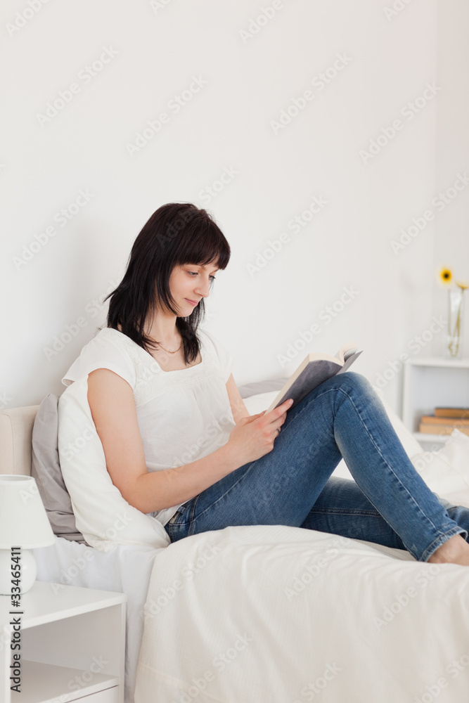 Charming brunette woman reading a book while sitting on a bed