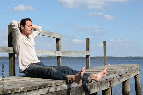 Fotografija Barefoot man sitting on a wooden jetty enjoying the sunshine