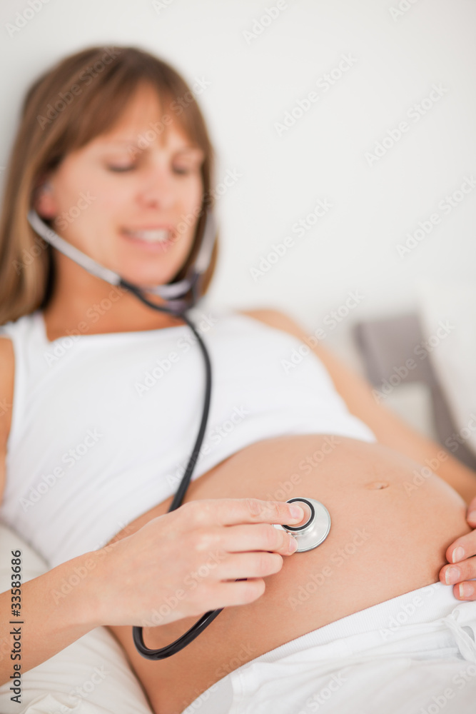 Attractive pregnant woman using a stethoscope while lying on a b Attractive pregnant woman using a stethoscope while lying on a b