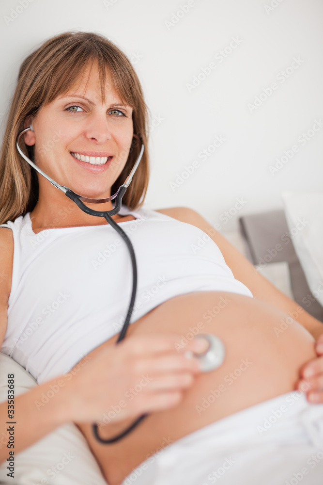 Nice pregnant woman using a stethoscope while lying on a bed Nice pregnant woman using a stethoscope while lying on a bed