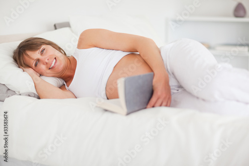 Cute pregnant woman reading a book while lying on her bed