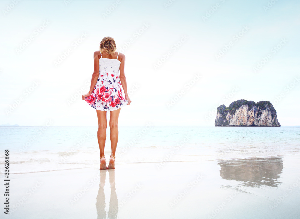 Young woman in summer dress standing on wet perfect sand and looking to her feet