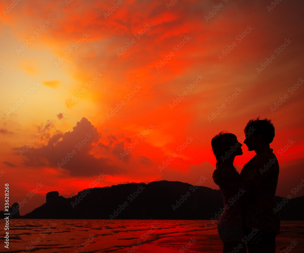 Young couple standing on the sand by sea on red dramatic sunset background