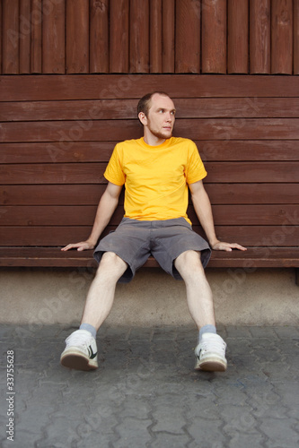 man in yellow shirt sitting on bench near wooden wall, looking a