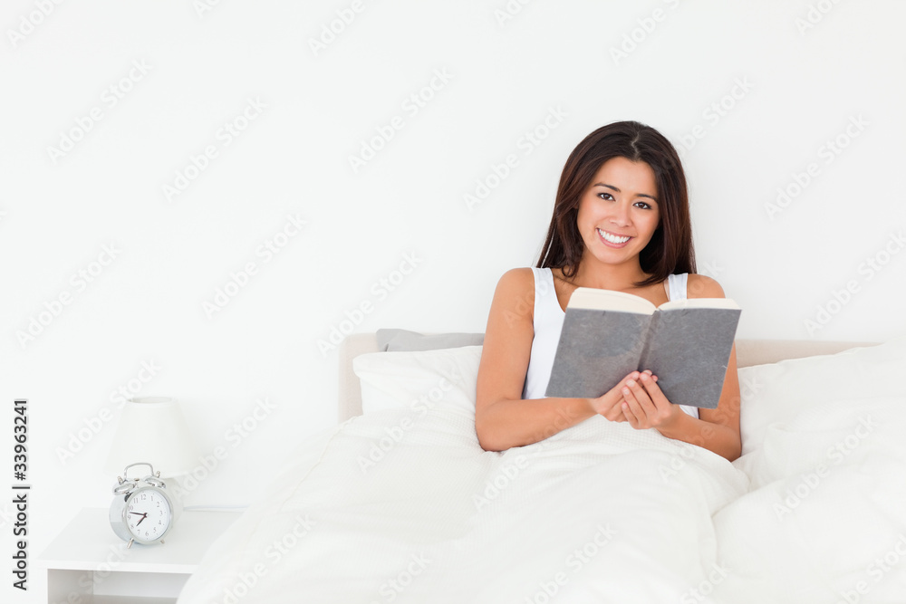 dark-haired woman holding book lyling in bed looking into camera dark-haired woman holding book lyling in bed looking into camera