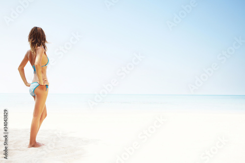 Young happy smiling woman in a swimsuit standing on a white-sand by the blue sea on clear blue sky background