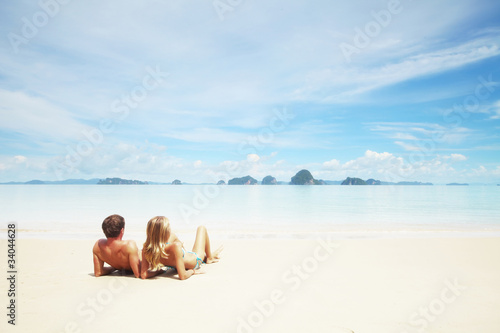 Young couple sitting on the sand and looking to a blue tropical sky