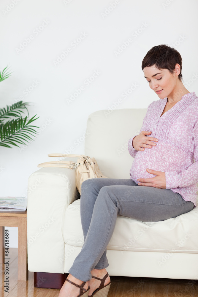 Smiling pregnant woman sitting on a sofa touching her belly Smiling pregnant woman sitting on a sofa touching her belly