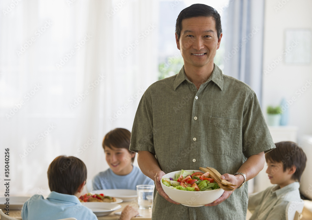 Smiling father holding salad with sons at table in background Smiling father holding salad with sons at table in background