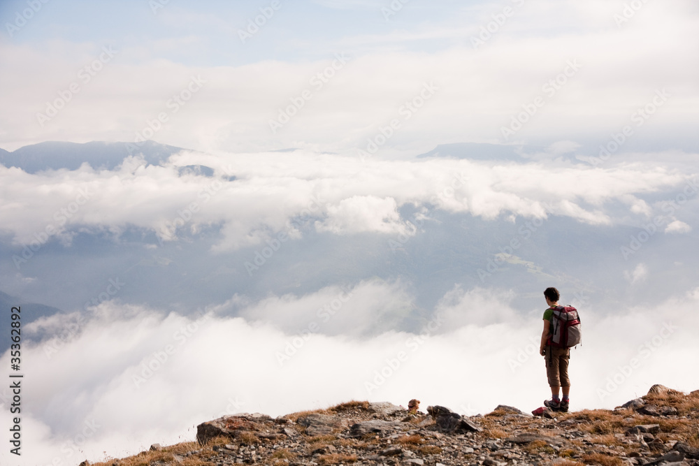 an adventurous man with a backpack alone on a mountain looking in the void and clouds in a very cold and cloudy day