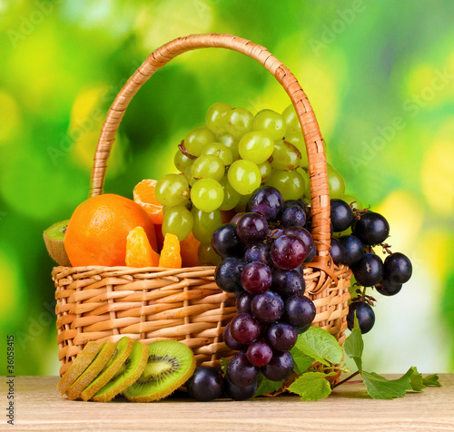 Ripe juicy fruits in basket on wooden table on green background