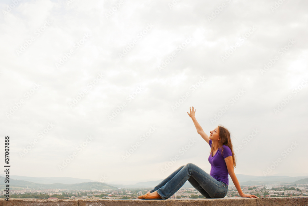 Young woman sitting with raised hand against blue sky Young woman sitting with raised hand against blue sky