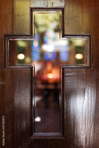 Cross shaped window in a door revealing the inside of a church.