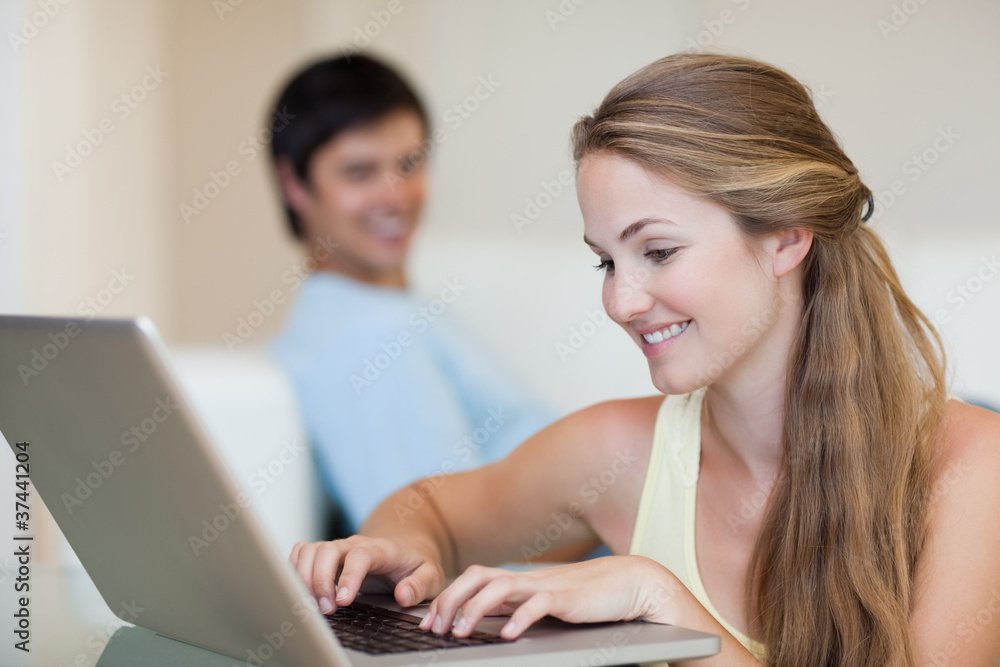 Woman using a notebook while her fiance is sitting on a couch Woman using a notebook while her fiance is sitting on a couch