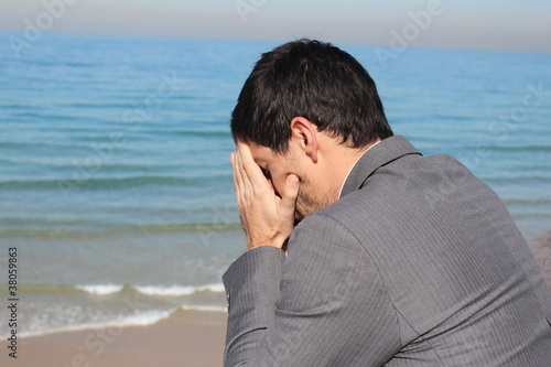 A businessman is holding his head, near the sea