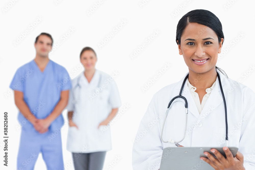 Smiling doctor with clipboard and staff behind her Smiling doctor with clipboard and staff behind her