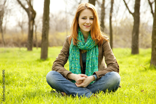 Beautiful red-haired girl sitting at green grass at park. © Masson