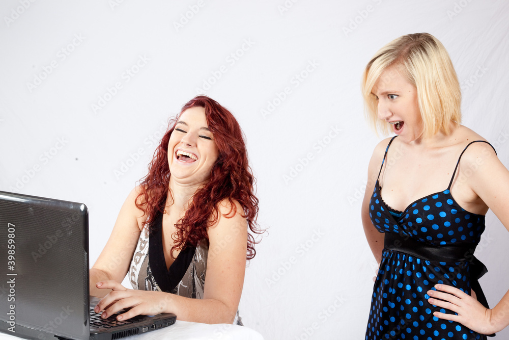 redhead woman laughing at a laptop while blond friend is shocked redhead woman laughing at a laptop while blond friend is shocked
