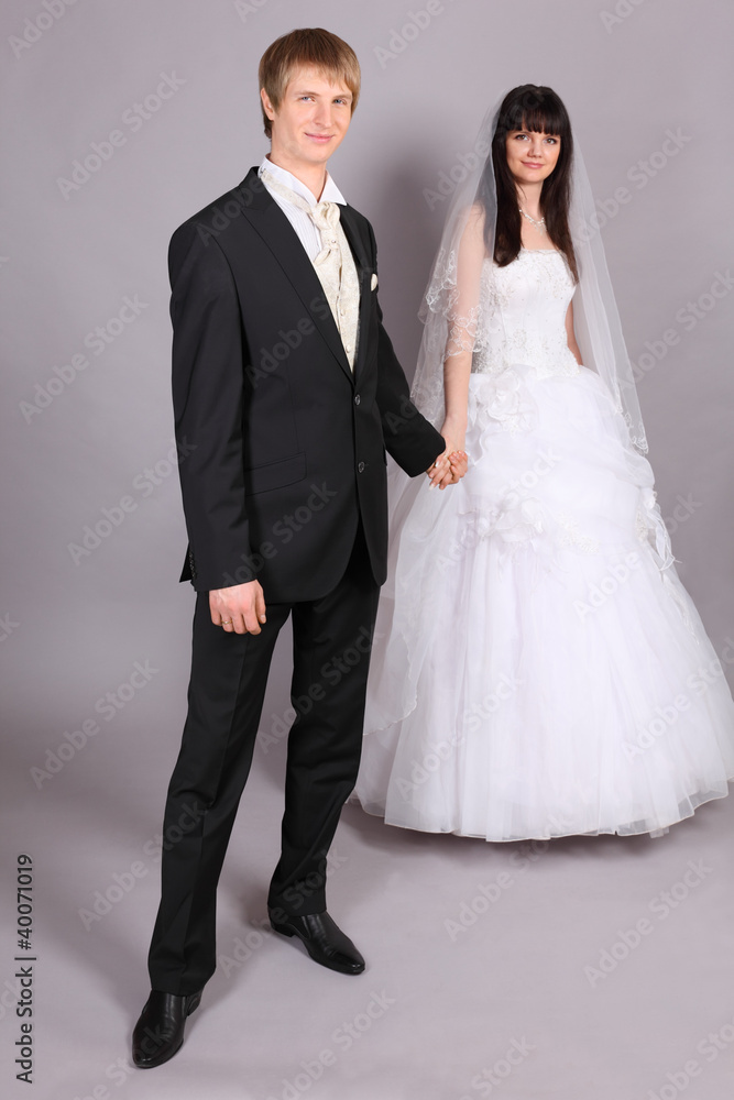 Beautiful bride and groom hold hands and stand in studio on gray