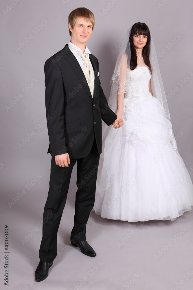Beautiful bride and groom hold hands and stand in studio on gray Beautiful bride and groom hold hands and stand in studio on gray