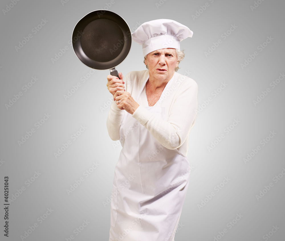 Cook senior woman angry trying to hit with pan over grey backgro Cook senior woman angry trying to hit with pan over grey backgro