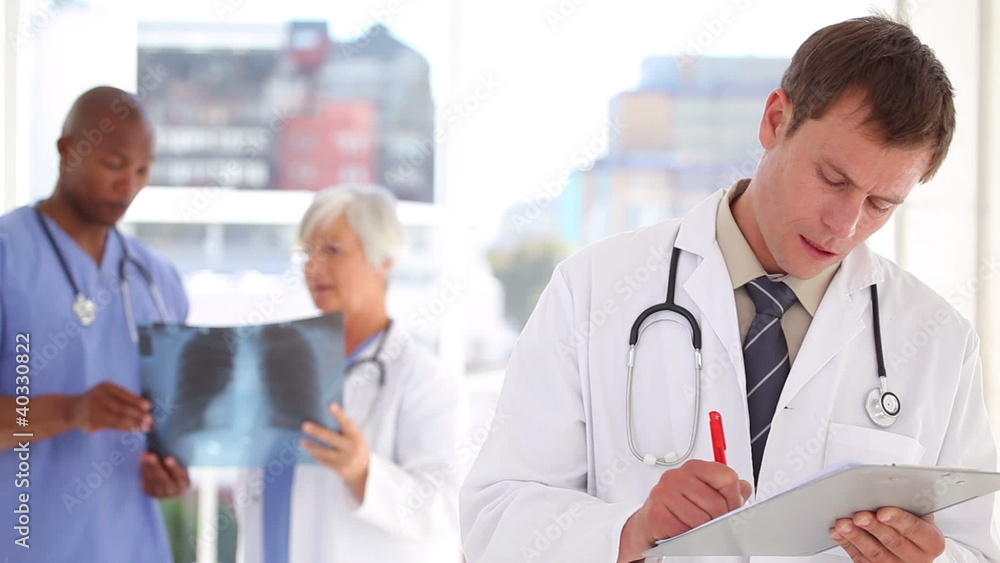 Serious doctor writing on his clipboard in front of colleagues Serious doctor writing on his clipboard in front of colleagues