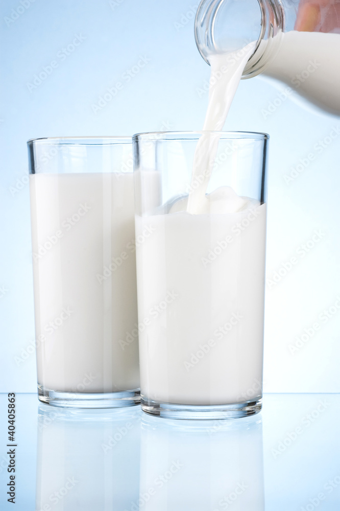 Pouring milk from a bottle into the two glasses on a blue backgr Pouring milk from a bottle into the two glasses on a blue backgr
