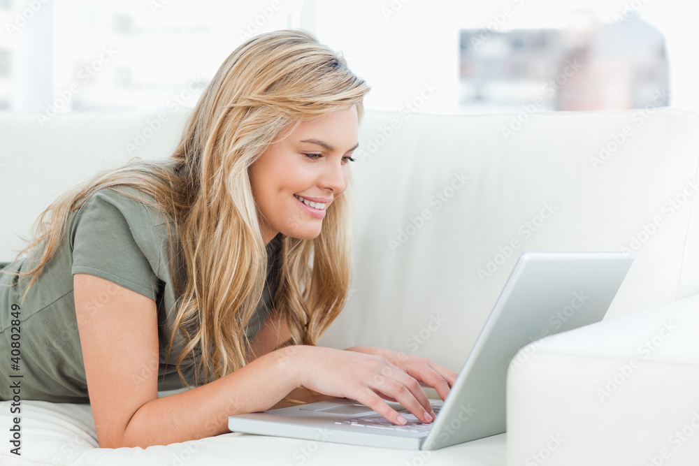 Woman smiling as she uses her laptop while lying across the couc Woman smiling as she uses her laptop while lying across the couc