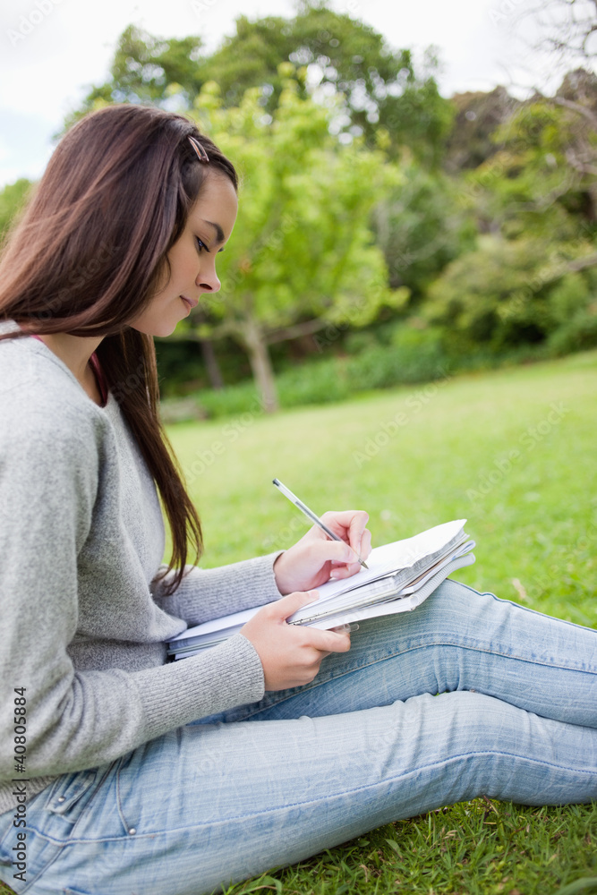 Obraz premium Side view of a young girl sitting on the grass while writing on