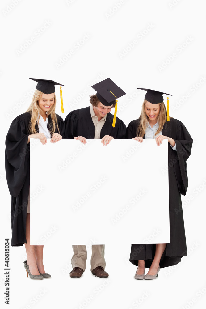Three smiling students in graduate robe holding a blank sign Three smiling students in graduate robe holding a blank sign